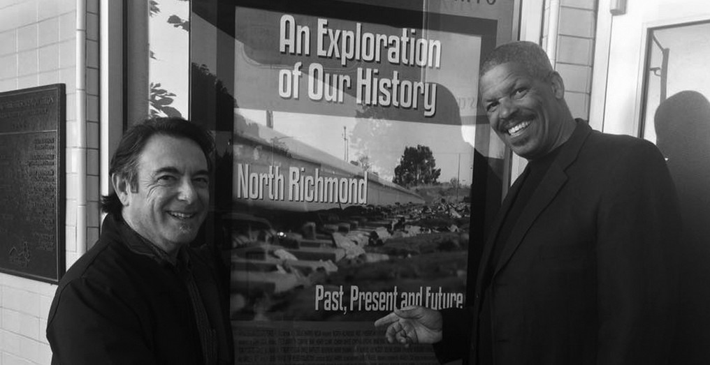 A black and white photo of Narrator John Gioia and Producer/Director Doug Harris at documentary premiere in 2016. They are standing in front of a poster of the exhibit that says "An Exploration of Our History: North Richmond - Past, Present, and Future."